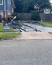 Worker spreading black tarp over driveway in front of house, with sign by lawn