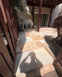 Covered stone patio with sunlit tiles, wooden posts, sliding glass doors, and wicker chairs.