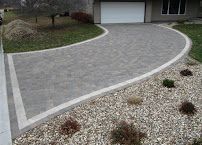 Curved gray paver driveway leading to a garage, bordered by light gravel and small shrubs