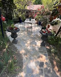 Sunlit stone patio garden with wrought-iron table, chairs, and lush potted flowers and greenery