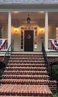 Front porch with brick steps, white columns, black railings, and a dark front door with wreath and warm lights