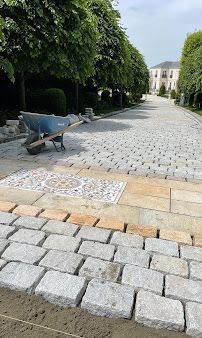 Cobblestone path with a wheelbarrow beside tall hedges and a building in the background