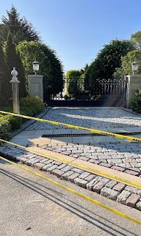 Cobblestone driveway sloping to a black iron gate, lined with yellow tape and stone pillars under a clear blue sky