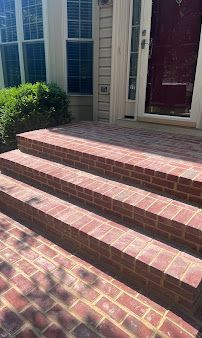 Brick steps leading to a house entrance with a white door and windows, sunlit with shadows.