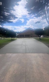 Concrete driveway leading to a tan building with a peaked roof, framed by trees and a cloudy blue sky.