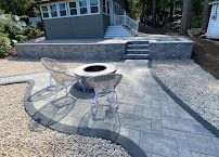 Backyard patio with two white chairs and a round fire pit on a stone paver patio in front of steps.