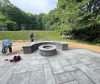 Outdoor stone patio with curved concrete bench and fire pit, workers installing the area near a grassy field and trees.