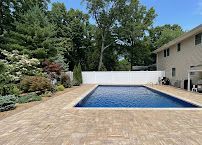 Backyard swimming pool with tan stone deck, white fence, and trees beside a beige house