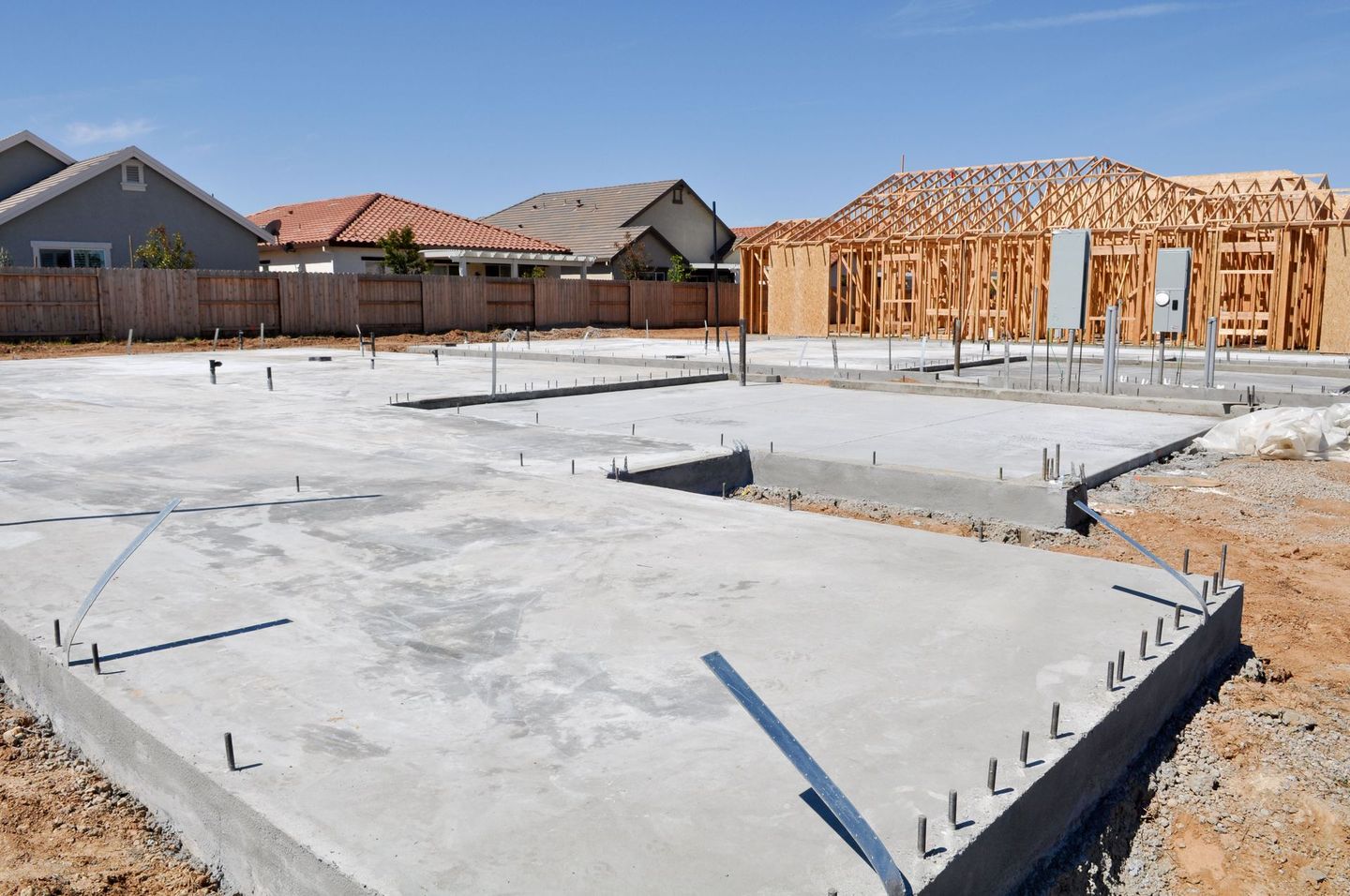 Concrete slab foundations laid for new residential homes in a suburban neighborhood, with one house in the framing stage and others in the background.