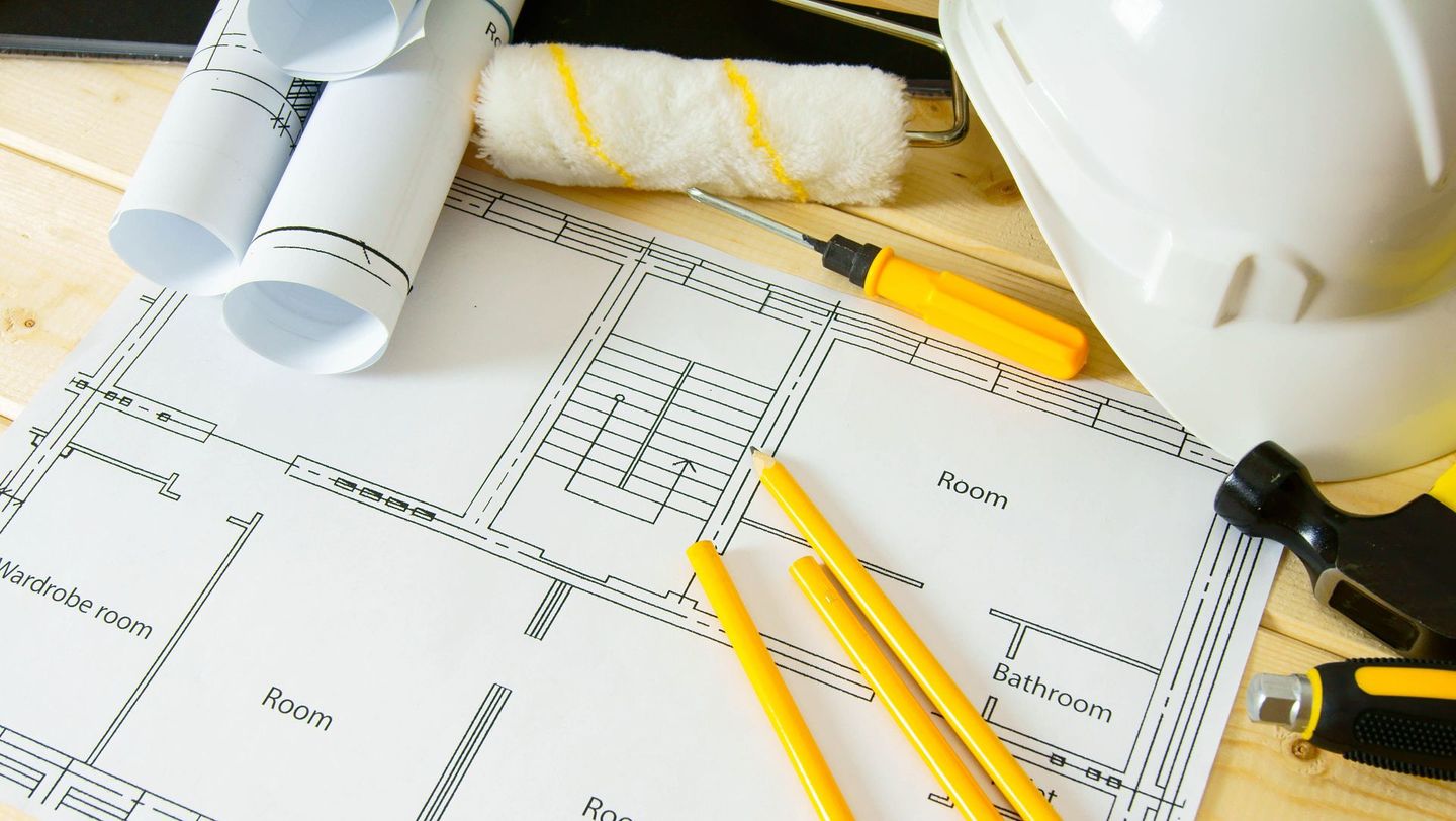 Architectural blueprints laid out on a wooden surface with construction tools, yellow pencils, a paint roller, and a white hard hat.