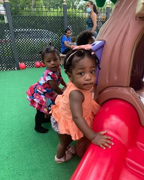 Children playing on green turf beside a red play structure in a fenced outdoor area.