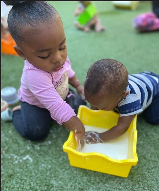 Two toddlers playing with water in a yellow tray on artificial grass.
