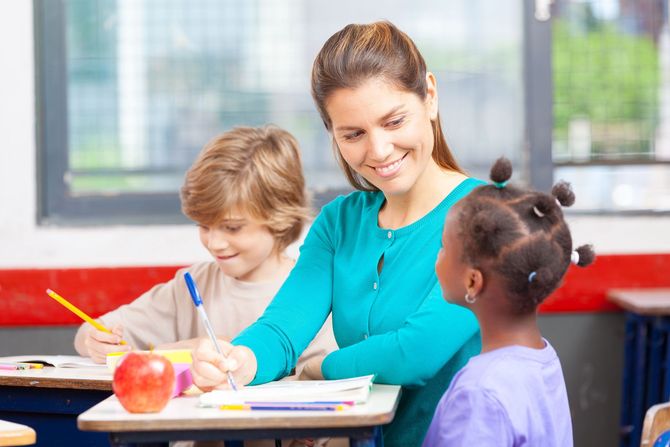 Teacher helping two children draw at a classroom table with crayons and paper