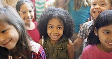 Children smiling and standing close together outdoors, with one child in a green jacket centered in front