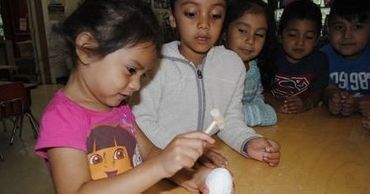 Children seated at a table, one girl in a pink shirt holding a white object while others watch.