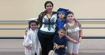 Five people pose together indoors, including one in a blue graduation cap and gown holding flowers.