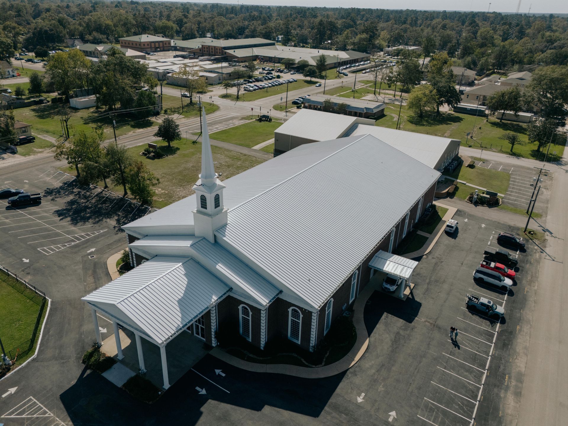Aerial view of a church with a tall steeple, gray siding, and parking lot, in a town.
