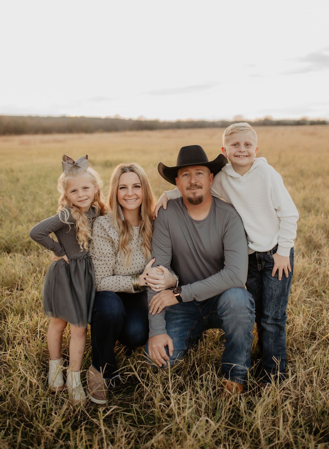 Family of four posing in a field at sunset: a man kneels, a woman stands, and two children smile.