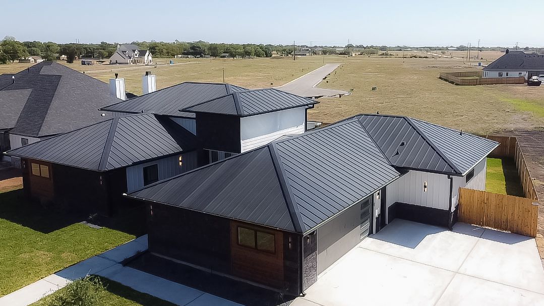 Aerial view of a modern house with a dark gray roof, concrete driveway, and surrounding green grass.