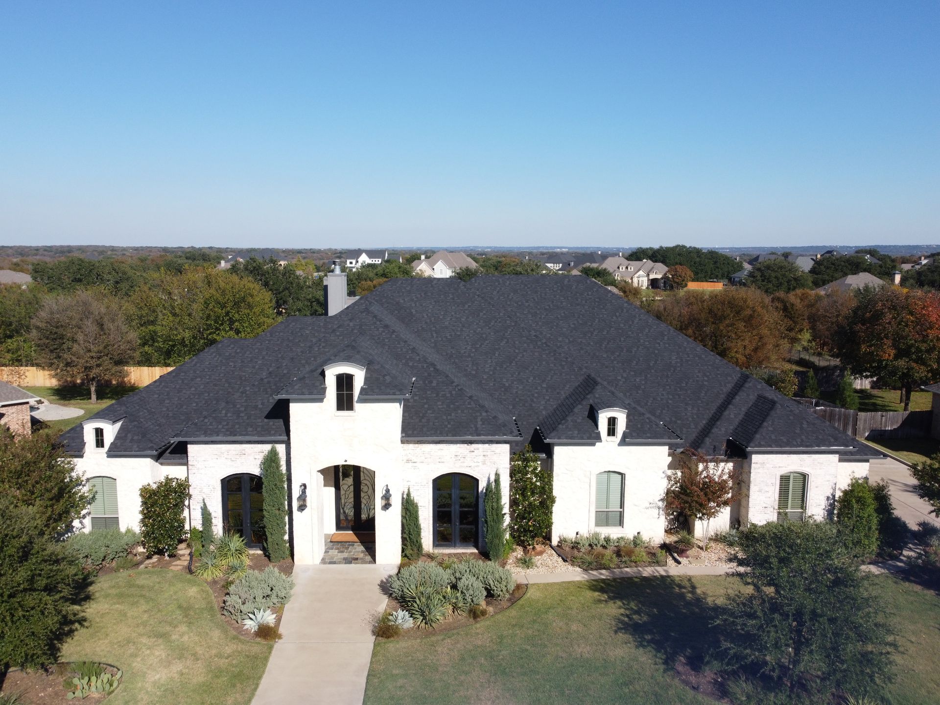 White brick house with black roof on a sunny day, surrounded by trees.