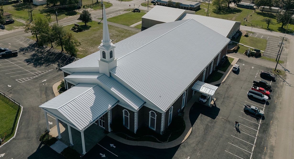 Aerial view of a church with a tall steeple, white metal roof, and parking lot