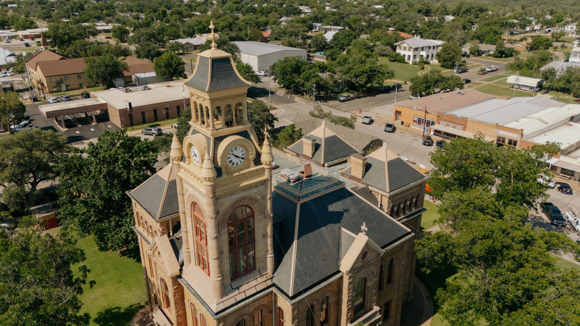 Aerial view of a clock tower with a light-colored facade, trees, and buildings in a small town.