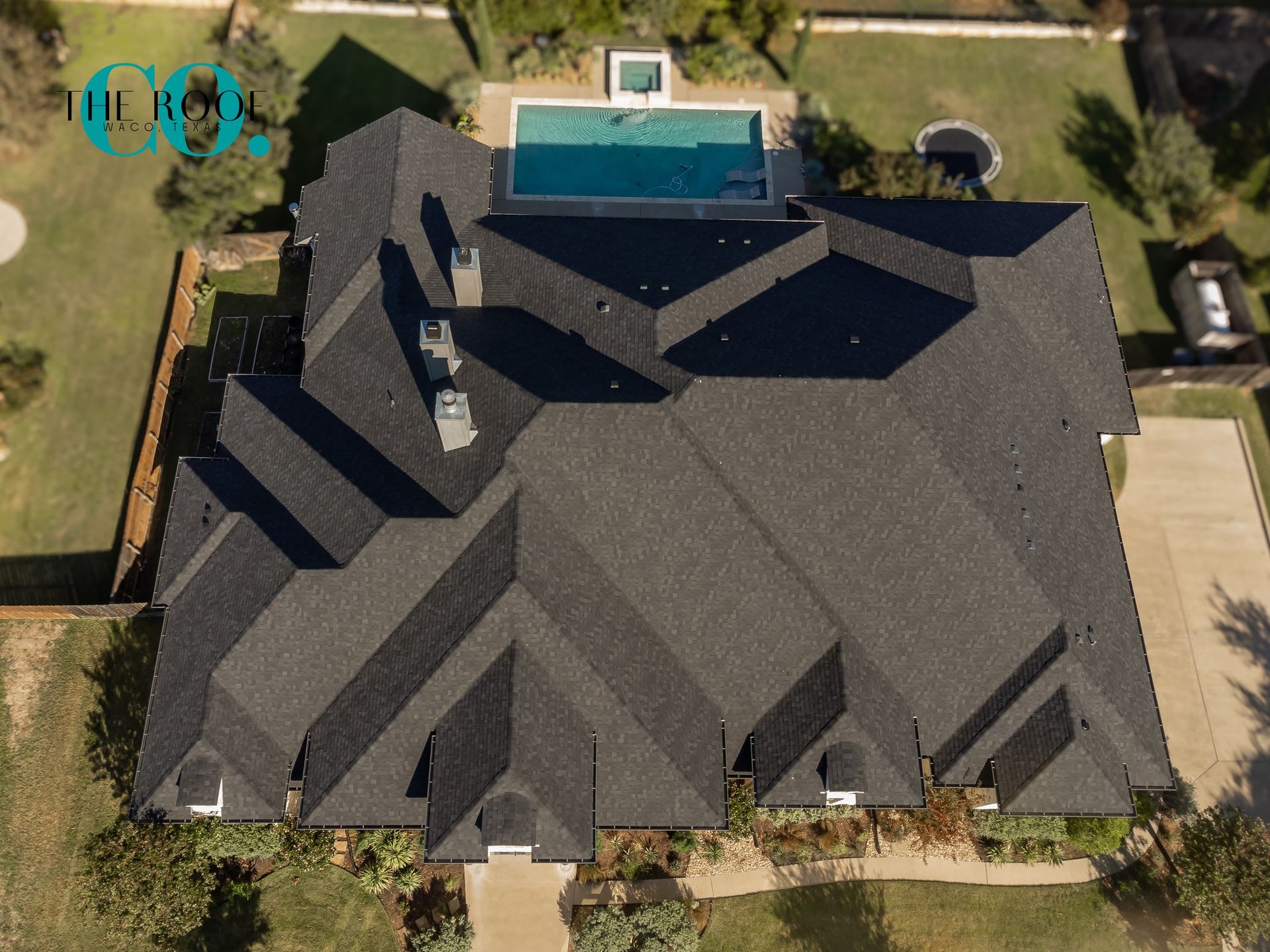 Aerial view of a dark-roofed house with a pool in the backyard, surrounded by green grass.