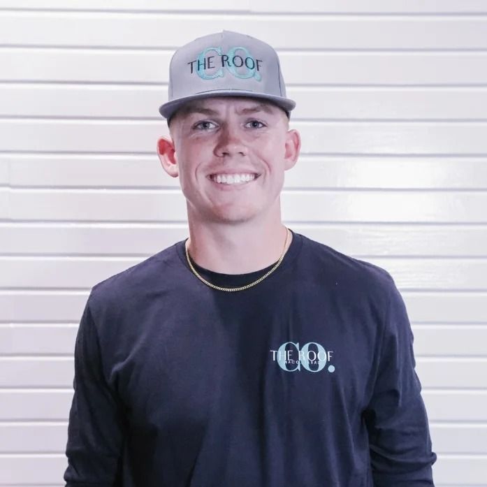 A young man wearing a gray baseball cap and a dark blue long-sleeved shirt, smiling at the camera. He is in front of a white, corrugated wall.