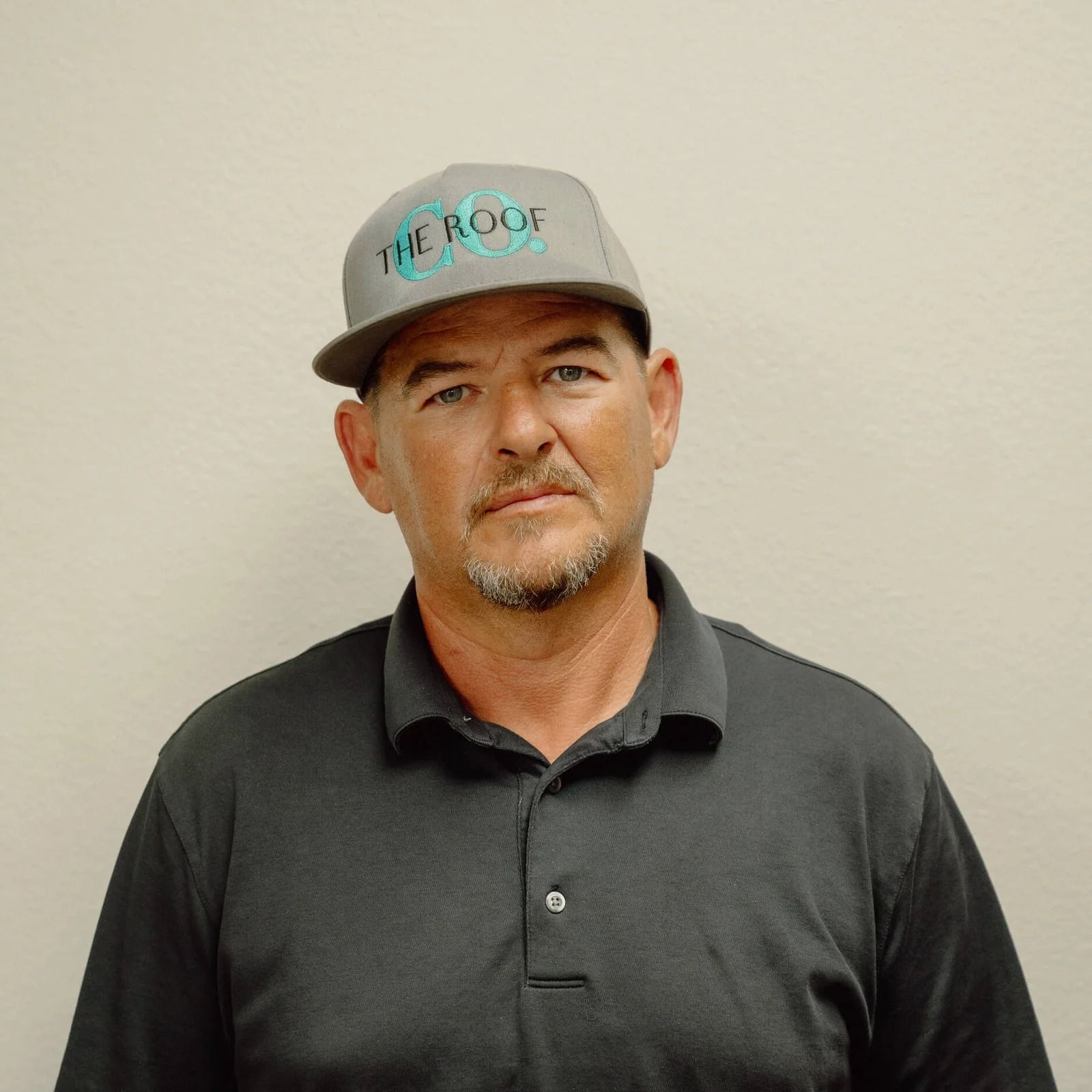 A man wearing a gray cap and shirt stands in front of a white wall. The cap reads 
