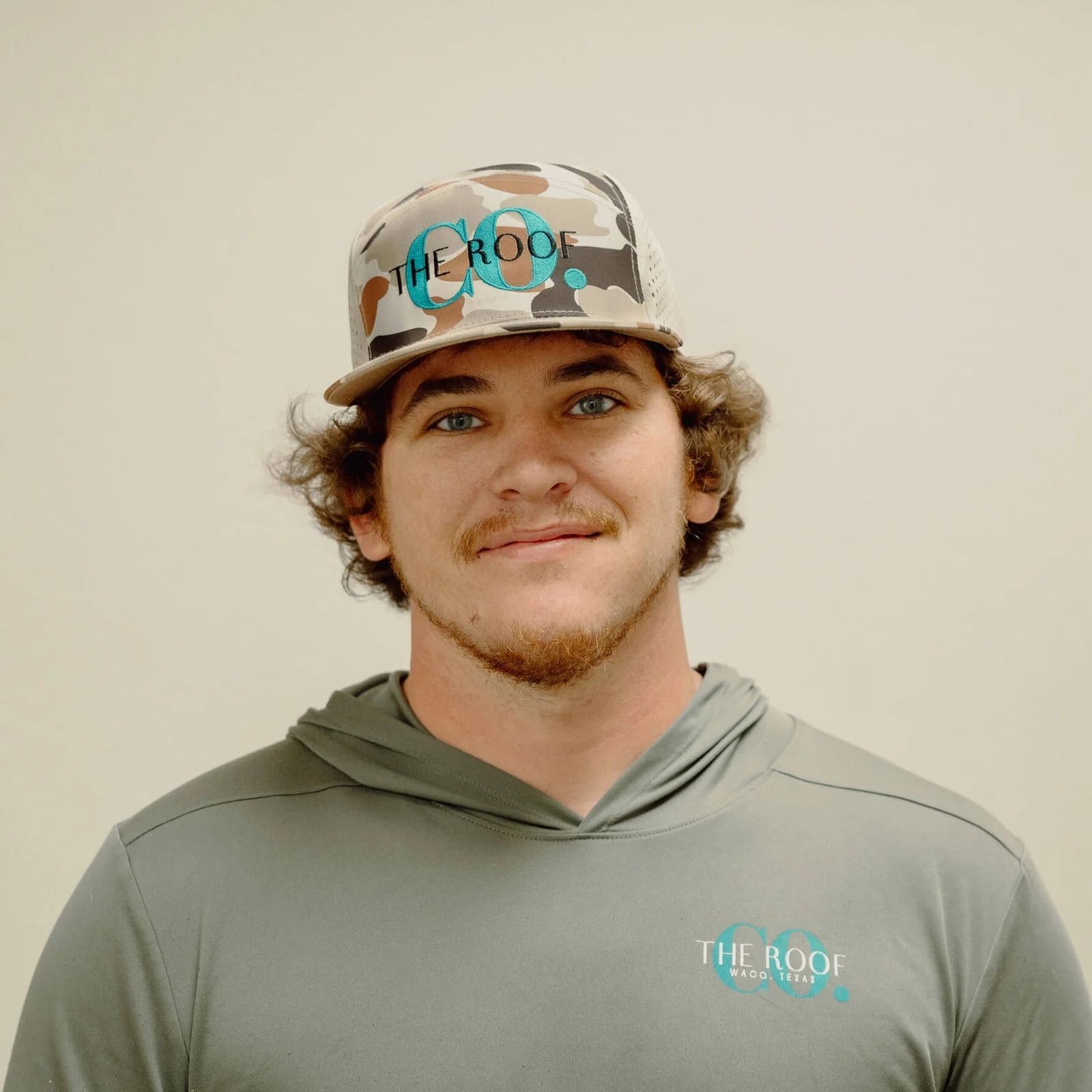 A young man with curly brown hair, wearing a camouflage hat and a gray hooded shirt, smiling.
