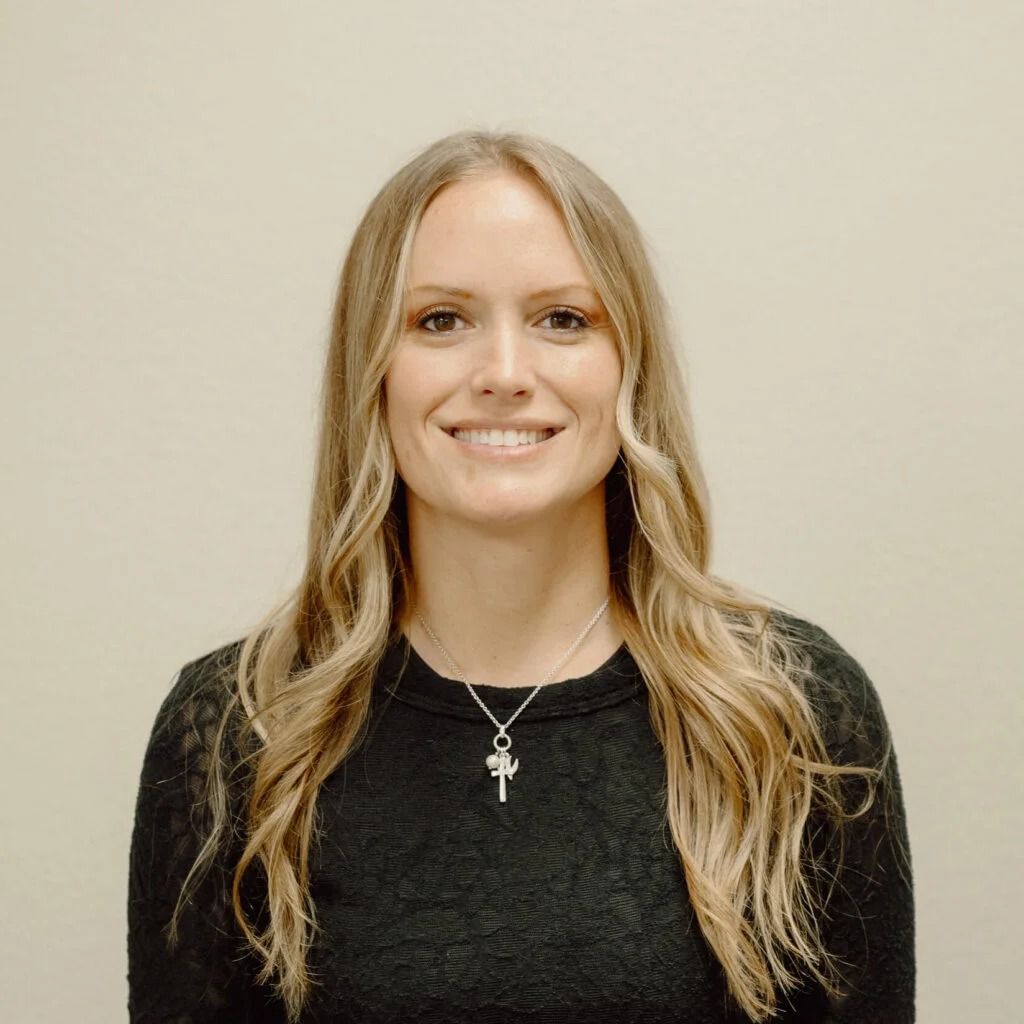 Woman with long blonde hair smiles at the camera, wearing a dark sweater and a silver pendant, set against a neutral wall.