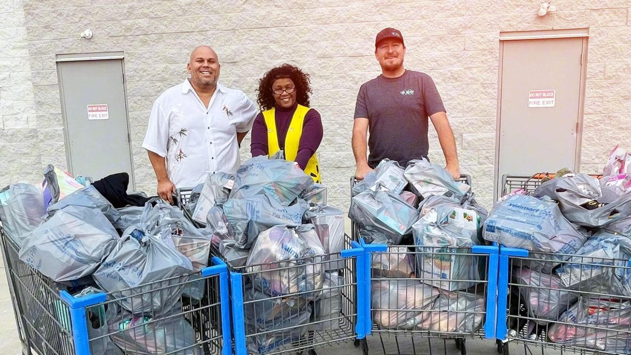 Three people stand behind shopping carts overflowing with bags of donations in front of a building. Two men and a woman smile.