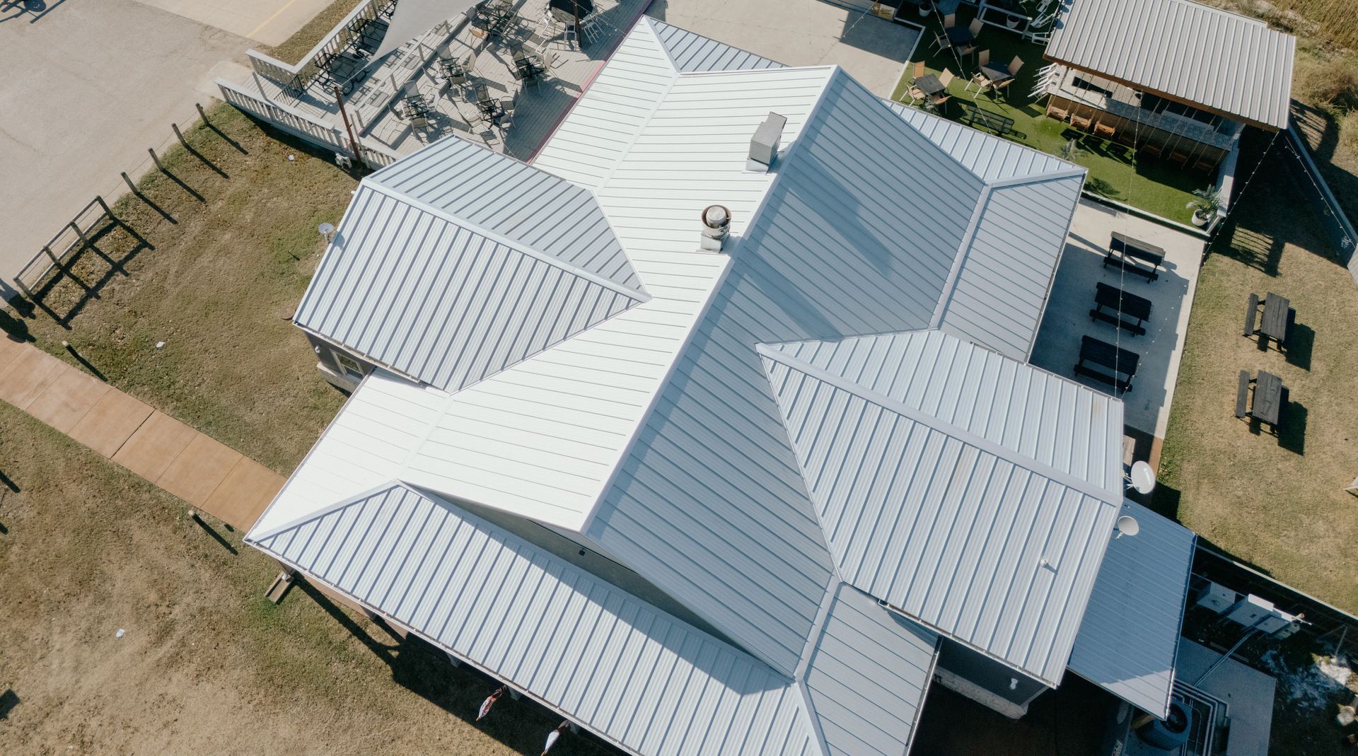 Aerial view of building with multi-faceted, white metal roof. Adjacent patio with seating.