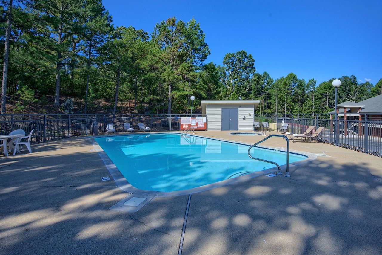 A rectangular outdoor pool with clear water, surrounded by concrete, a fence, and trees under a blue sky.