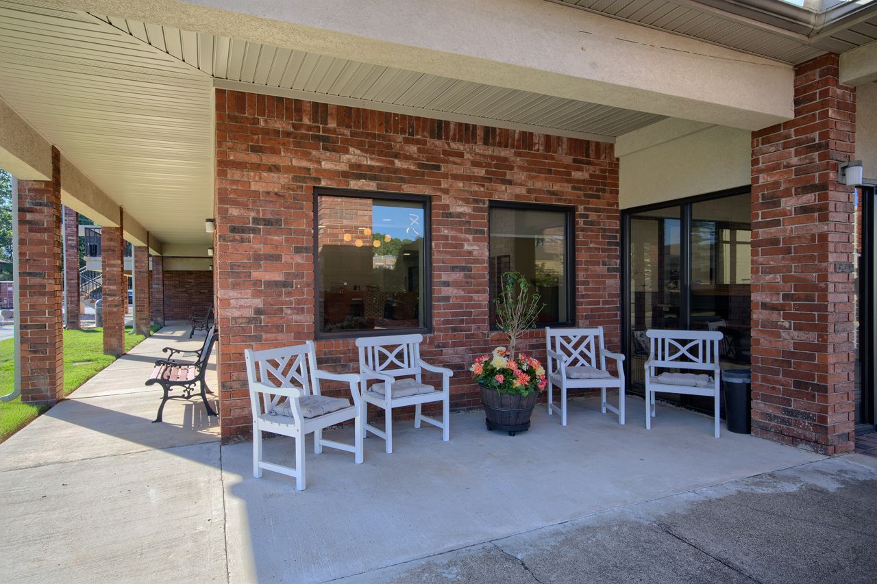 Covered porch with white chairs, brick wall, potted flowers, and windows.