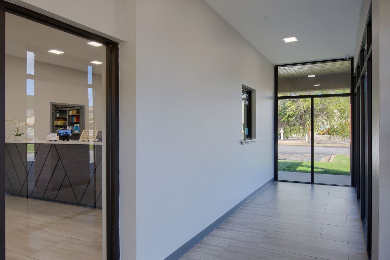 Hallway with glass-walled entrance and reception desk visible through a glass door. Grey walls, dark trim, and a concrete floor.
