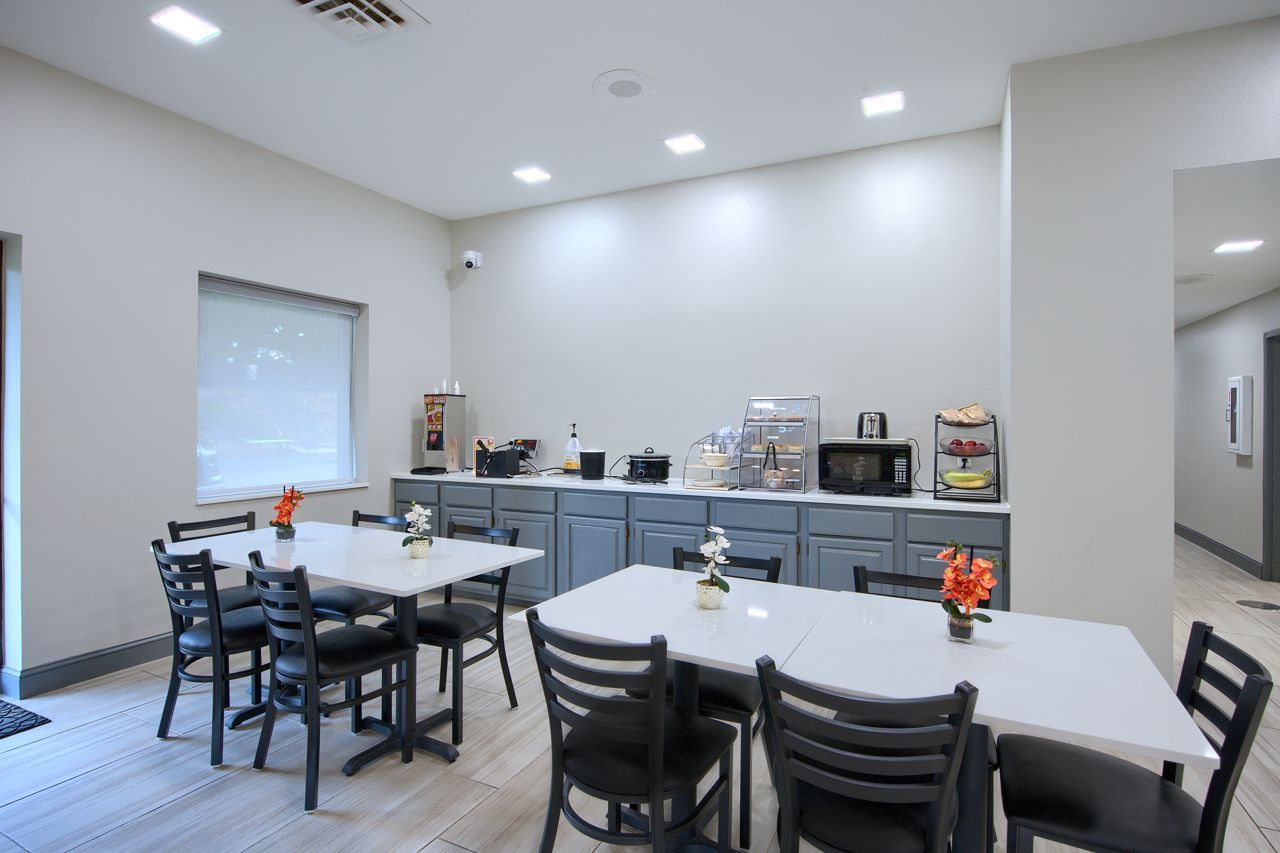 Hotel breakfast area with tables, chairs, and buffet. Countertop appliances and a TV are visible.