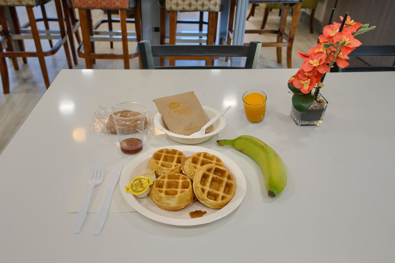 Breakfast spread: waffles, banana, juice, muffin, fruit, and flowers on a white table.