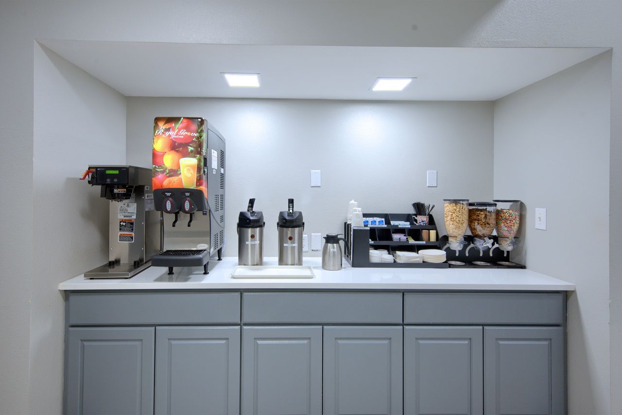 Breakfast station with coffee maker, juice dispenser, cereal, and toppings, set against a neutral gray wall.