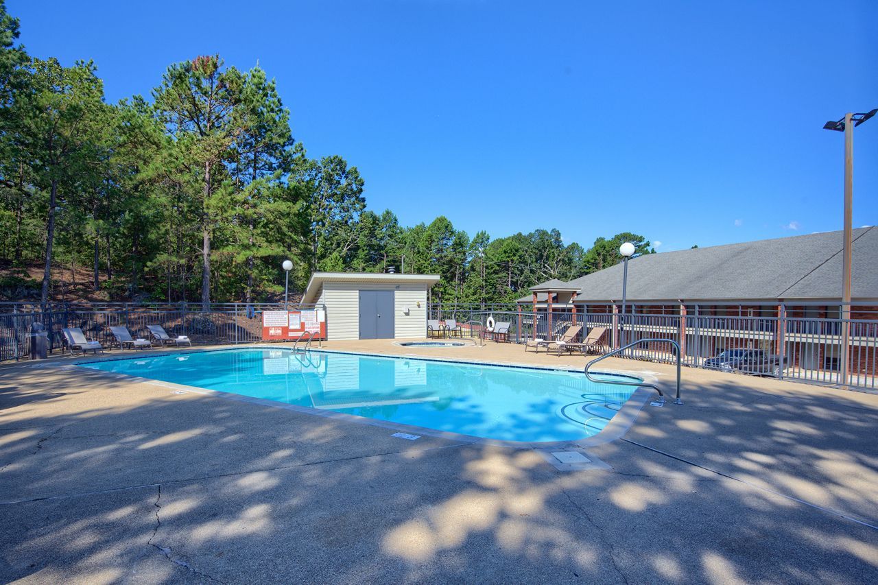Pool surrounded by concrete and trees, near a building with a dark roof under a clear blue sky.