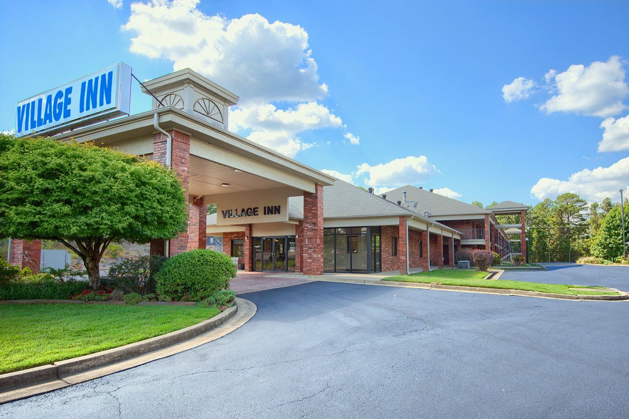 Village Inn hotel with red brick facade and blue signage. Driveway leads to entrance under a covered area, sunny day.