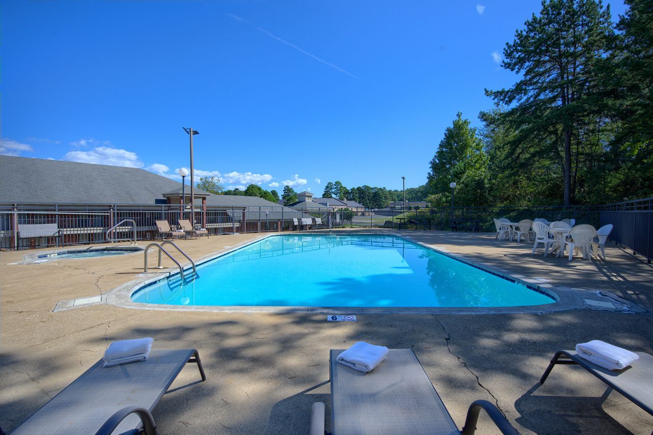 Swimming pool with lounge chairs, blue water, and clear sky. Building and trees in the background.