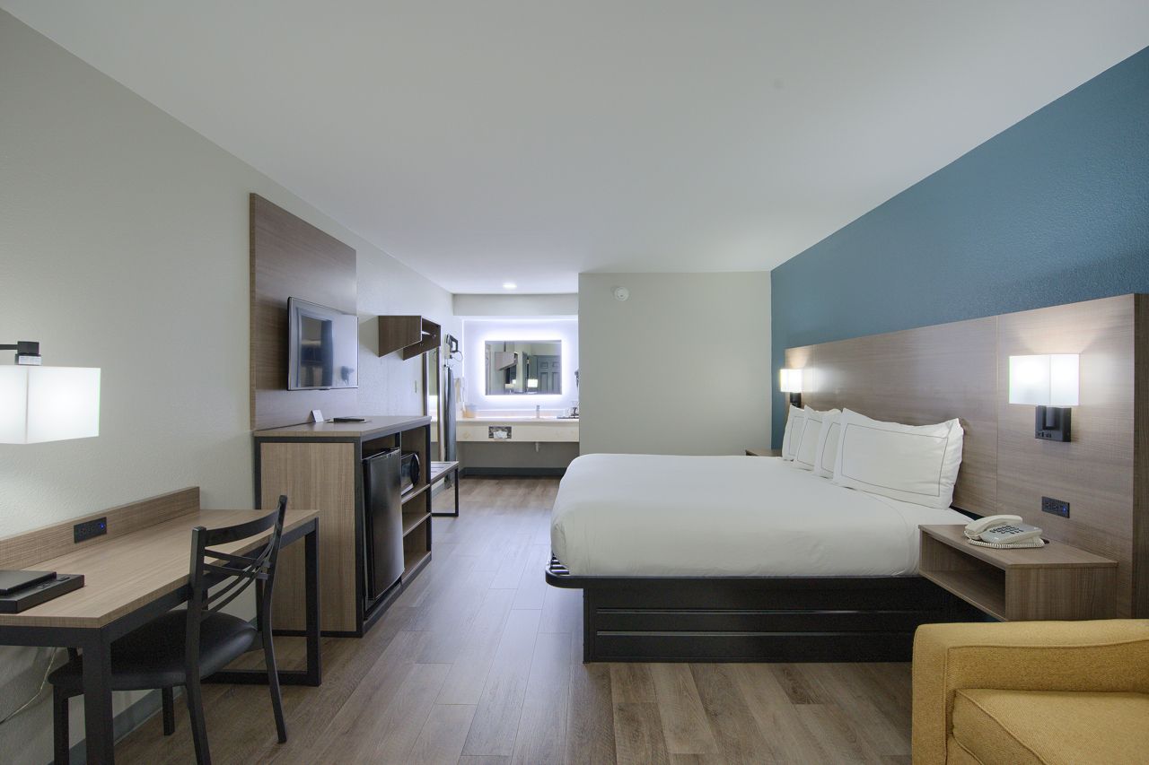 Hotel room with bed, desk, kitchenette, and bathroom visible. Neutral colors.