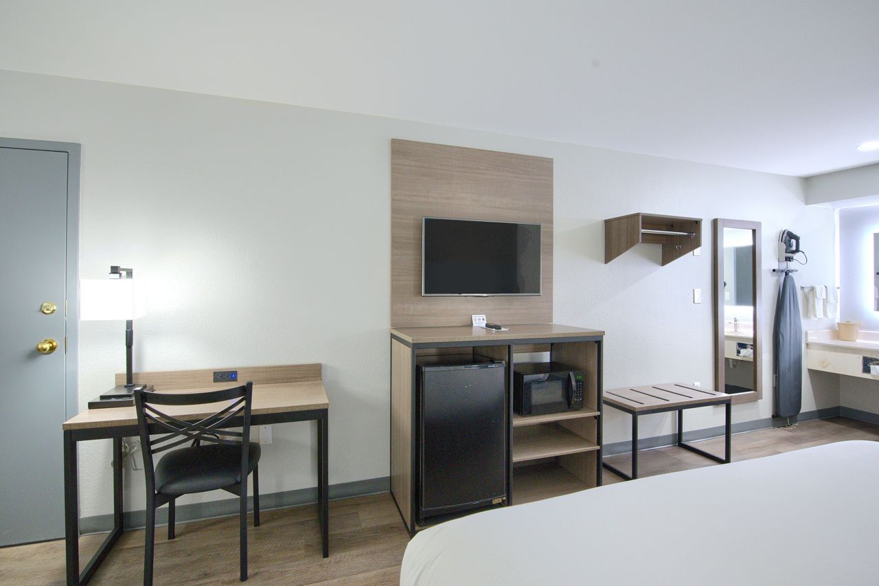 Hotel room with desk, TV, fridge, and bed. Light walls, gray door.