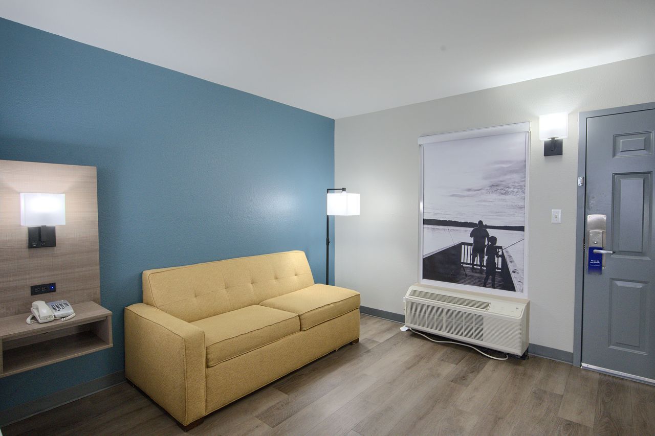 Hotel room interior: yellow sofa, blue wall, bedside table, window with a scene, and a closed door.