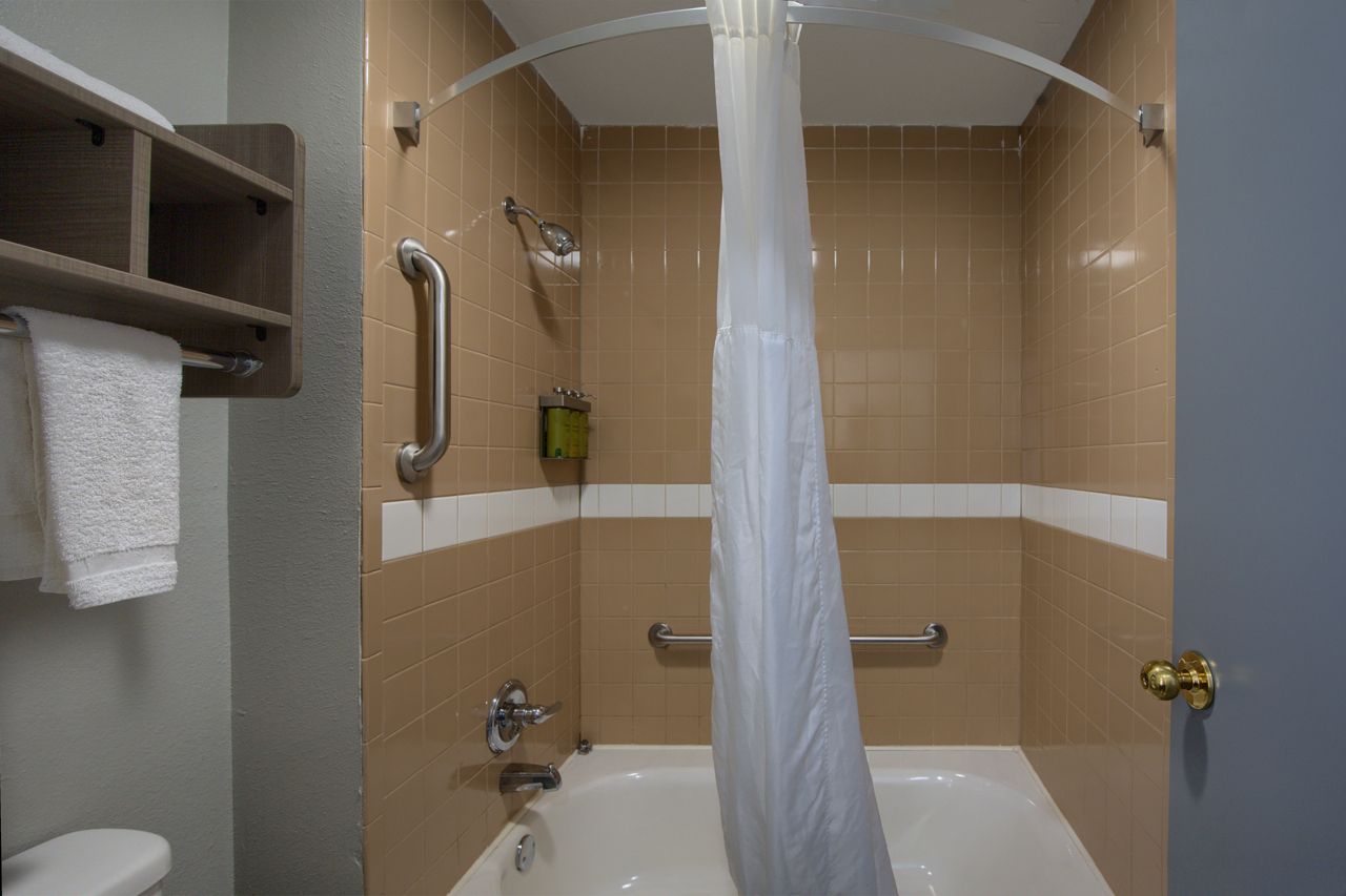 Bathroom with a tub, shower curtain, grab bars, and shelf with towels. Beige tiles and a gray wall.