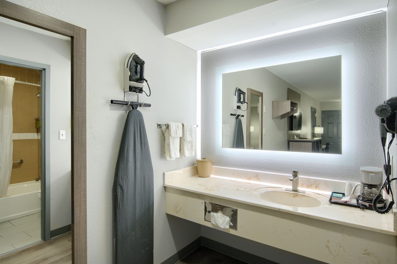 Hotel bathroom with illuminated mirror, vanity, iron, and doorway to the shower.
