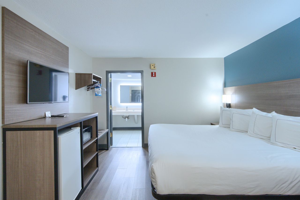 Hotel room with king-sized bed, TV on wall, mini-fridge, and bathroom visible in the doorway.
