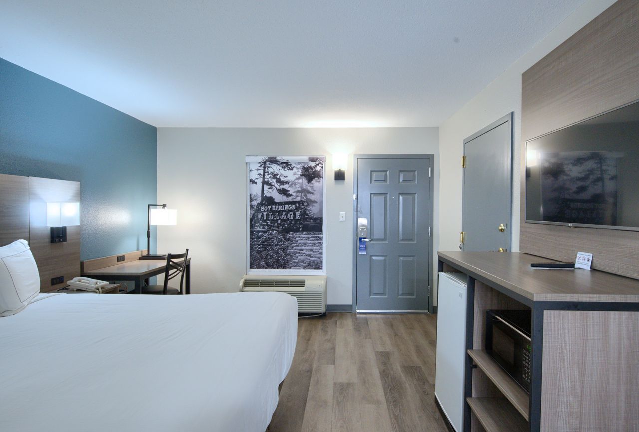 Hotel room with a bed, desk, TV, and door; neutral colors.