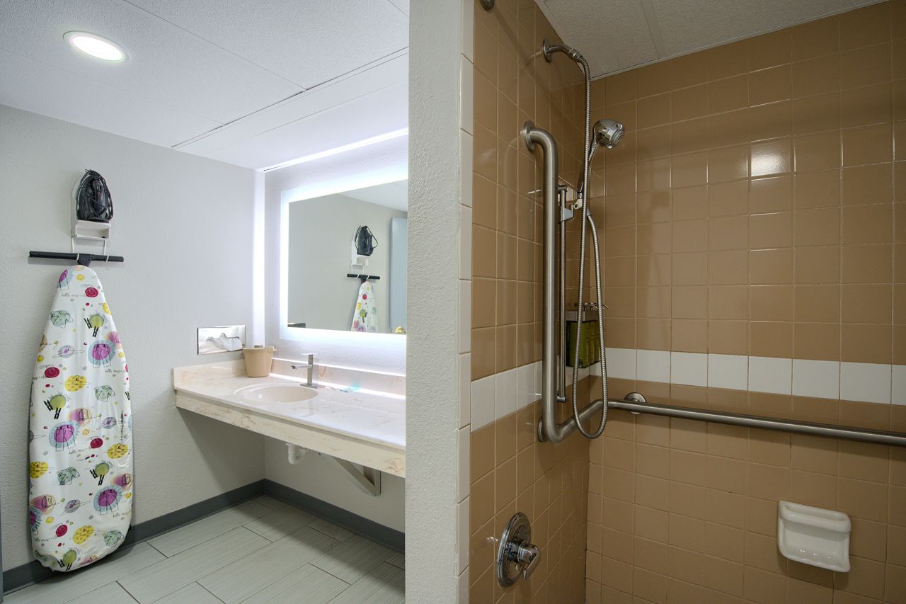 Bathroom with shower, vanity, and ironing board. Beige and white tiled walls, with a mirror and shower fixtures.